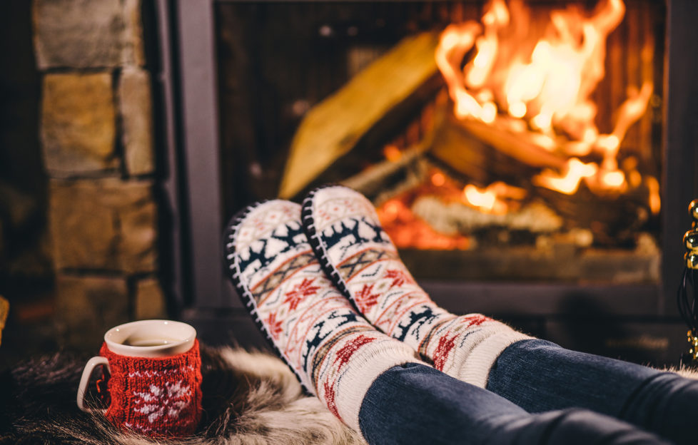 Feet in woollen socks by the Christmas fireplace. Woman relaxes by warm fire with a cup of hot drink and warming up her feet in woollen socks. Close up. Winter and Christmas holidays concept. ;