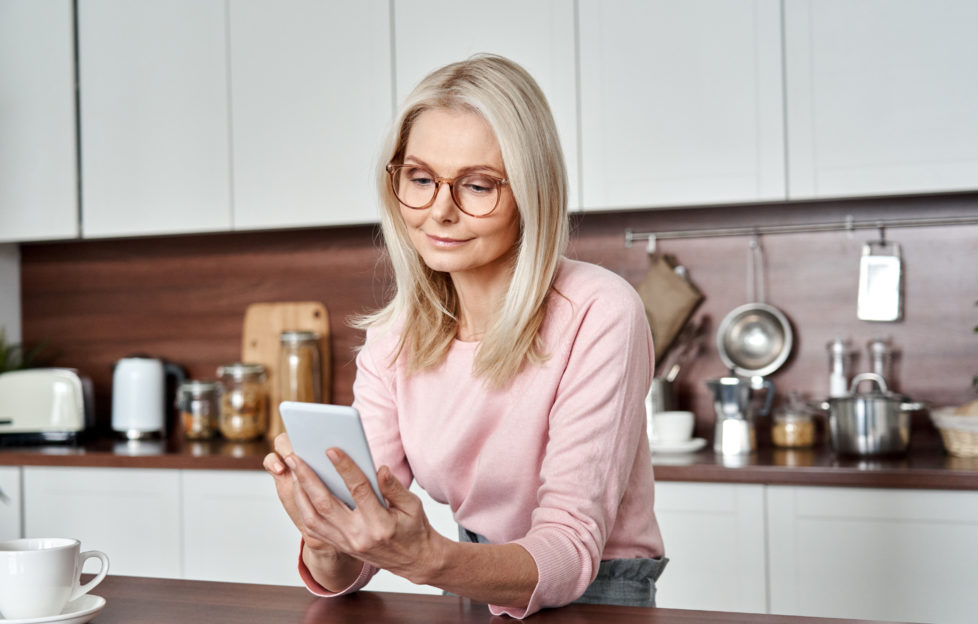Middle aged 50 years old woman using apps ordering buying food on smartphone sitting in kitchen at home. Mature older lady holding mobile phone texting messages, browsing online services.;