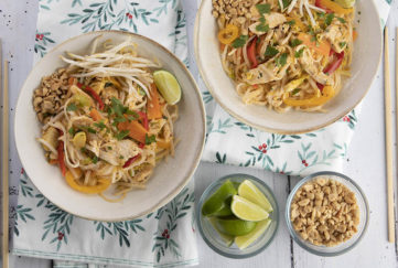 Two bowls of chicken pad thai sit on a tablecloth decorated with a foliage pattern. There are small bowls of limes and peanuts beside them.