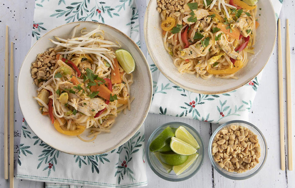 Two bowls of chicken pad thai sit on a tablecloth decorated with a foliage pattern. There are small bowls of limes and peanuts beside them.