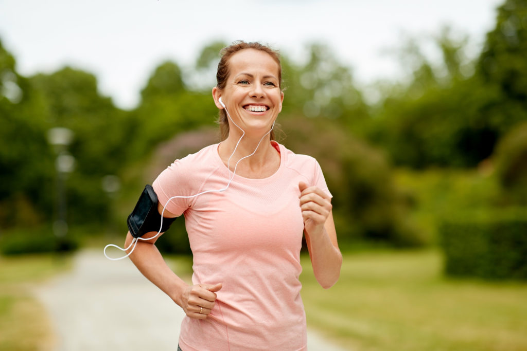 fitness, sport and healthy lifestyle concept - smiling woman with earphones wearing armband for smartphone, jogging at summer park and listening to music; 