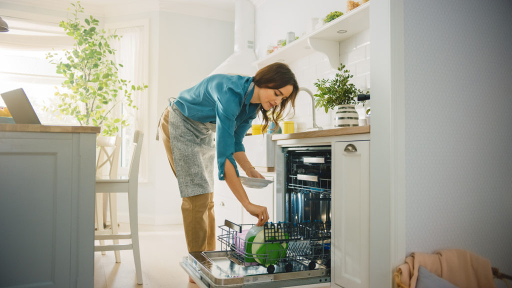Beautiful Female is Loading Dirty Plates into a Dishwasher Machine in a Bright Sunny Kitchen. Girl in Wearing an Apron. Young Housewife Uses Modern Appliance to Keep the Home Clean.; 