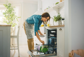 Beautiful Female is Loading Dirty Plates into a Dishwasher Machine in a Bright Sunny Kitchen. Girl in Wearing an Apron. Young Housewife Uses Modern Appliance to Keep the Home Clean.;