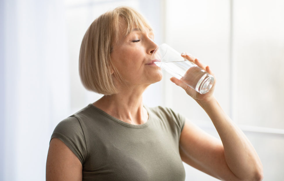 Fit senior woman drinking clear water during her workout break at home. Mature Caucasian lady staying hydrated after sports training. Healthy lifestyle and wellness concept;