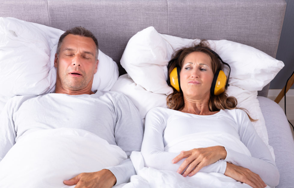 Mature Woman Covering Her Ears With Headphone While Man Snoring In Bed;