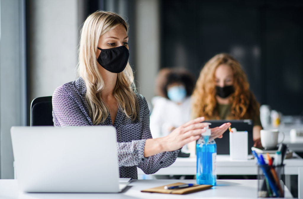 Young woman with face mask back at work in office after lockdown, disinfecting hands.