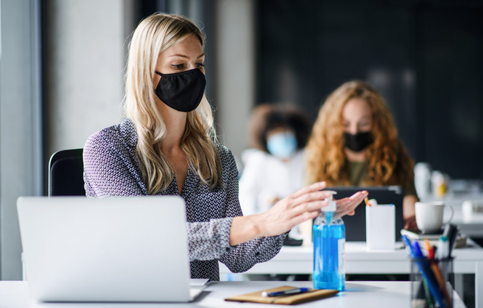 Young woman with face mask back at work in office after lockdown, disinfecting hands.