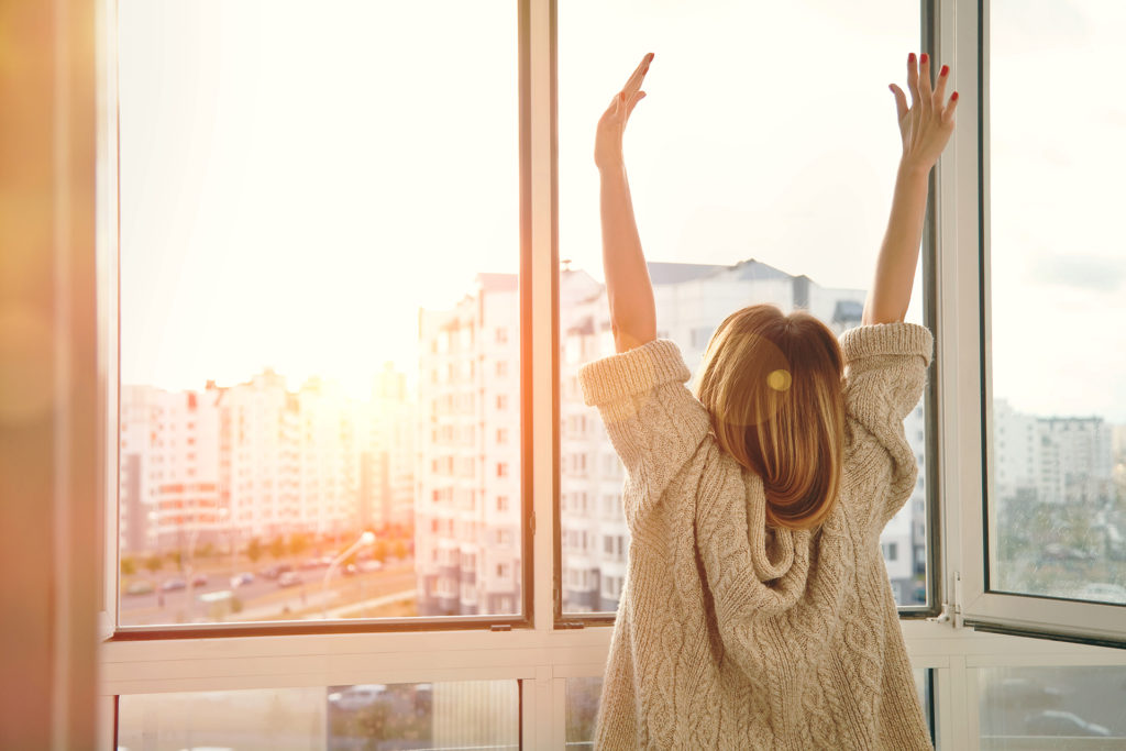 Woman near window raising hands facing the sunrise at morning; 