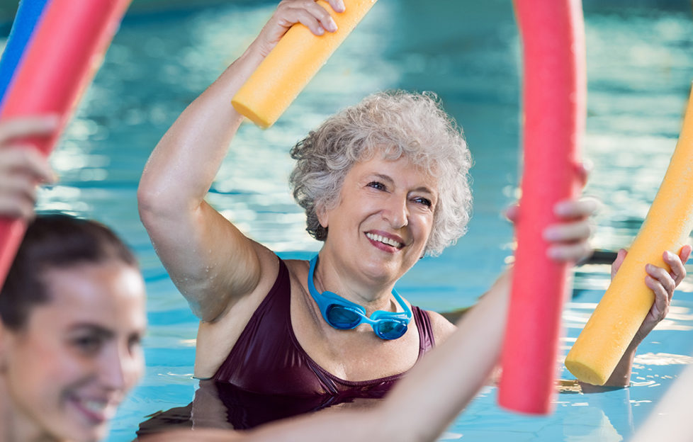 Smiling senior woman doing aqua fitness with swim noodles. Happy mature healthy woman taking fitness classes in aqua aerobics. Healthy old woman holding swim noodles doing aqua gym with young trainer.;