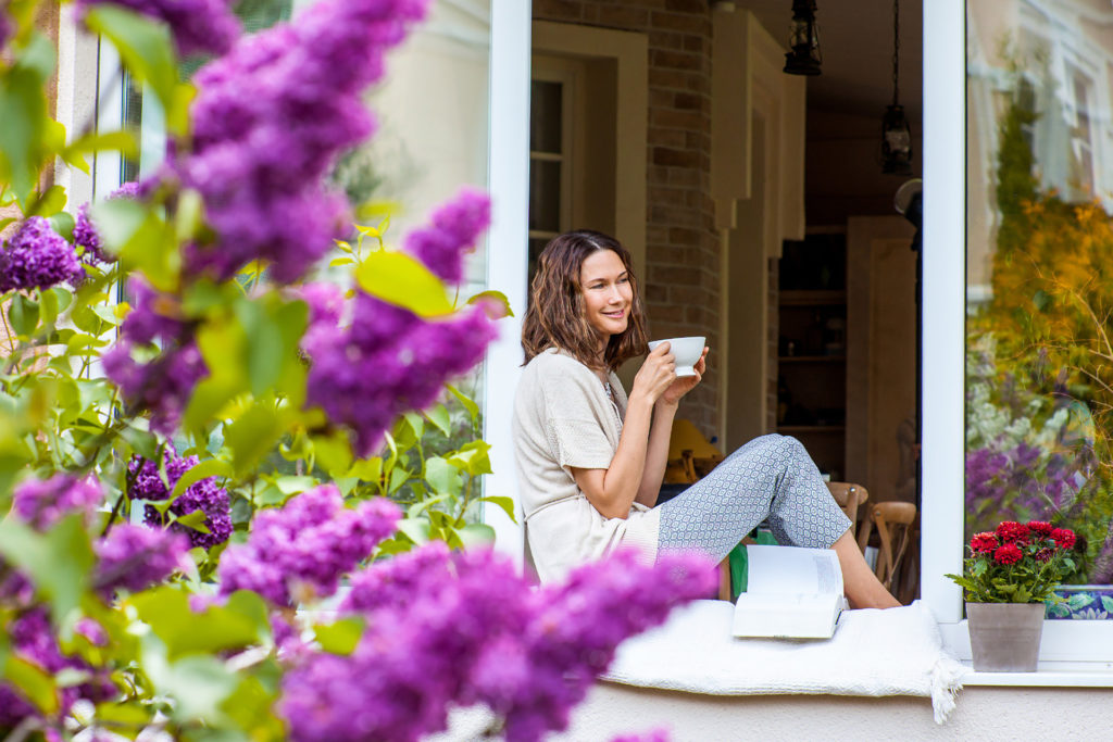 Smiling beautiful middle-aged woman with a cup of hot drink on the windowsill in the garden with blooming lilac; 