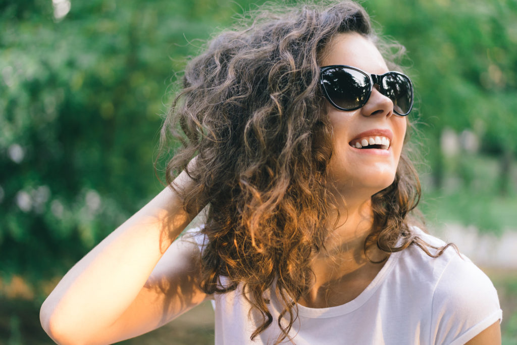 Portrait of a young smiling happy woman in sunglasses at the park on a background of green trees. Girl with curly hair outdoors in the summer.