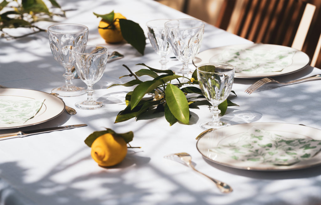 Outdoor table set with white cloth, antique cutlery, china plates and fresh lemons with leaves