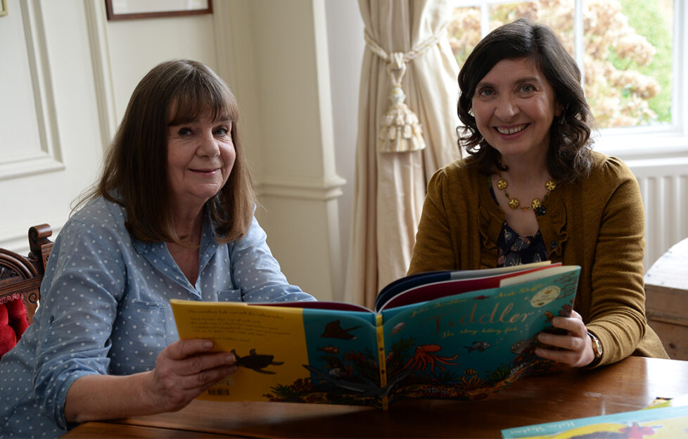 2 mature women looking at a large children's picture book, smiling