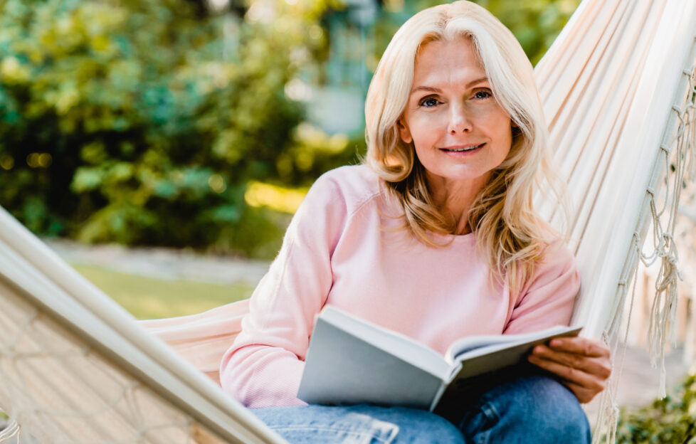 Nice-looking mature blonde woman resting in hammock in the garden with a book;