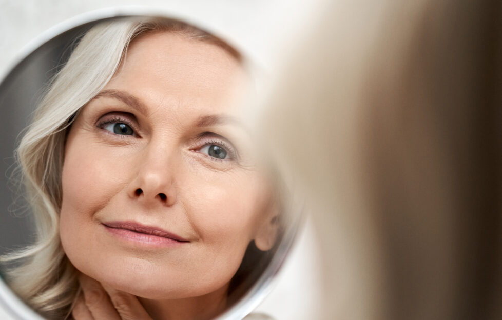 Happy 50s middle aged woman model touching face skin looking in mirror reflection. Smiling mature old lady pampering, healthy moisturized skin care, aging beauty, skincare treatment cosmetics concept.;