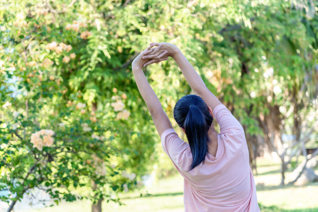 Back view of young fitness woman doing stretch exercise outdoors in park. Asian female stretching her arms above head for warming up before running or working out in morning. 