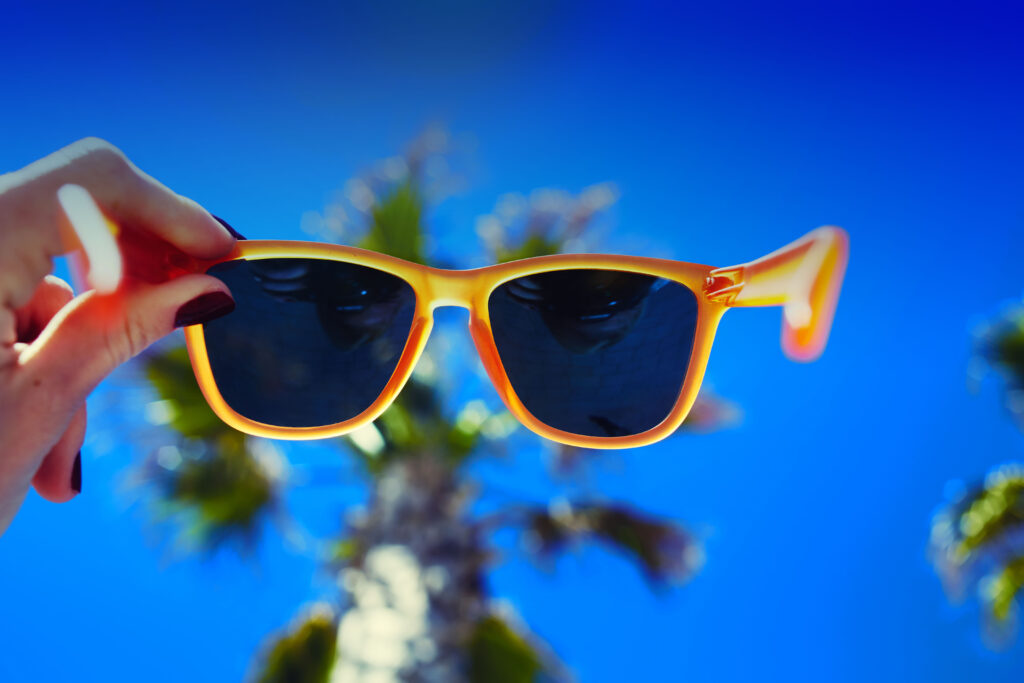 Female hand holding colorful sunglasses against palm tree and blue sunny sky, summer vacation holidays concept, first person shot, looking though glasses; 