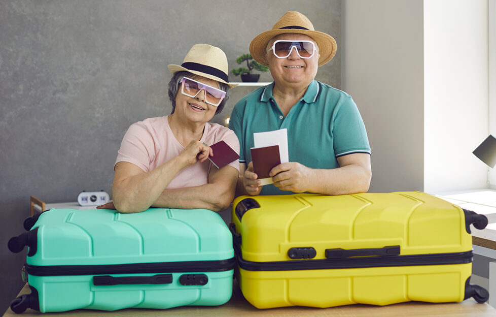 Mature husband and wife at home before going on family holiday. Portrait of happy cute active senior couple with flight tickets, passports and suitcases standing in living-room and smiling