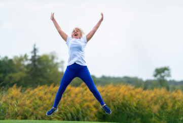 Older woman wearing workout outfit jumping for joy with arms and legs spread in midair in front of blurry yellow flowers