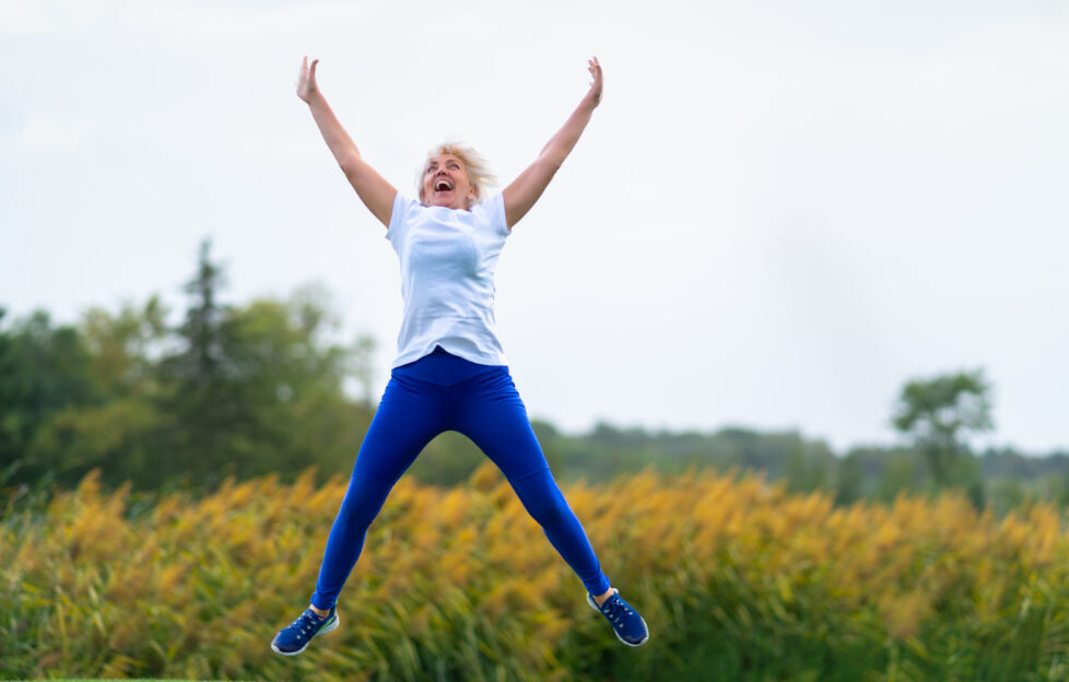 Older woman wearing workout outfit jumping for joy with arms and legs spread in midair in front of blurry yellow flowers