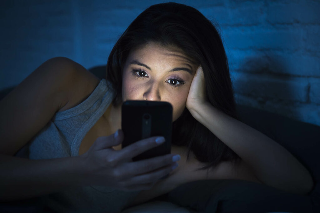 young beautiful womanl in bed using mobile phone late at night at dark bedroom lying happy and relaxed enjoying social media network at her phone in communication internet addiction concept;