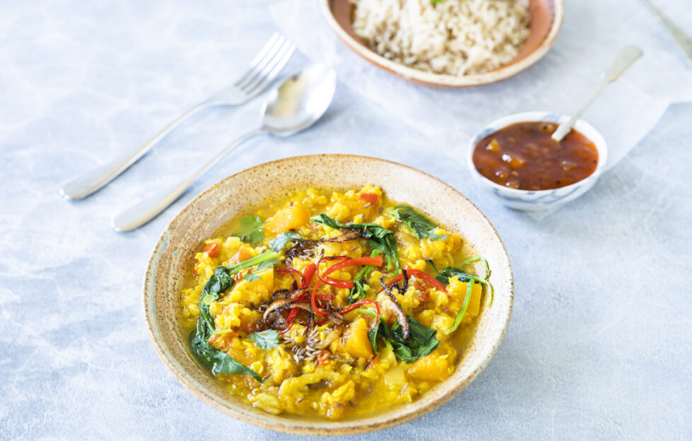 A bowl of yellow dal curry sits on a blue tablecloth beside a knife and fork and accompaniments of rice and pickle.