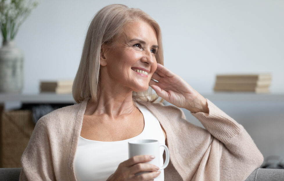 Happy beautiful dreamy old senior middle aged woman sit on sofa relax at home hold cup drink coffee tea looking away thinking dreaming enjoy stress free peaceful mood wellbeing alone in living room;