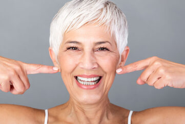 Beautiful Caucasian smiling senior woman with short grey hair pointing at her teeth and looking at camera. Beauty photography.;