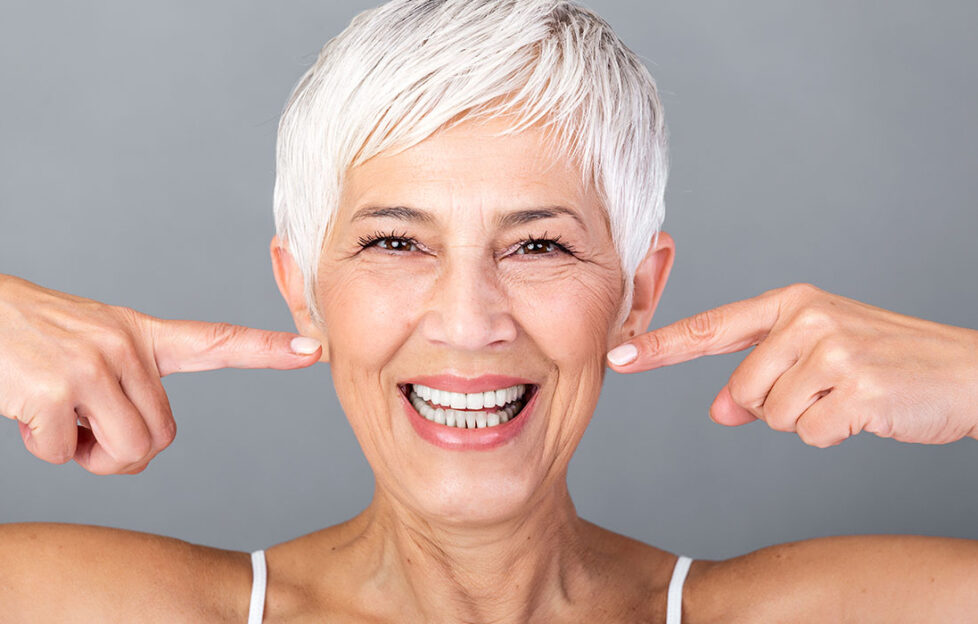 Beautiful Caucasian smiling senior woman with short grey hair pointing at her teeth and looking at camera. Beauty photography.;