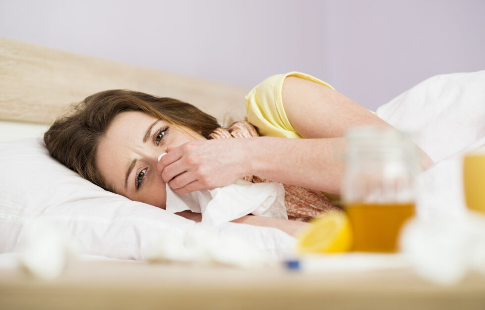 Sick woman lying in bed with high fever. She has cold and flu. In front of her is tea with lemon and honey.;