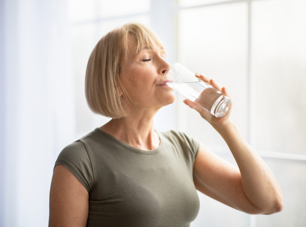 Fit senior woman drinking clear water during her workout break at home. Mature Caucasian lady staying hydrated after sports training. Healthy lifestyle and wellness concept; 