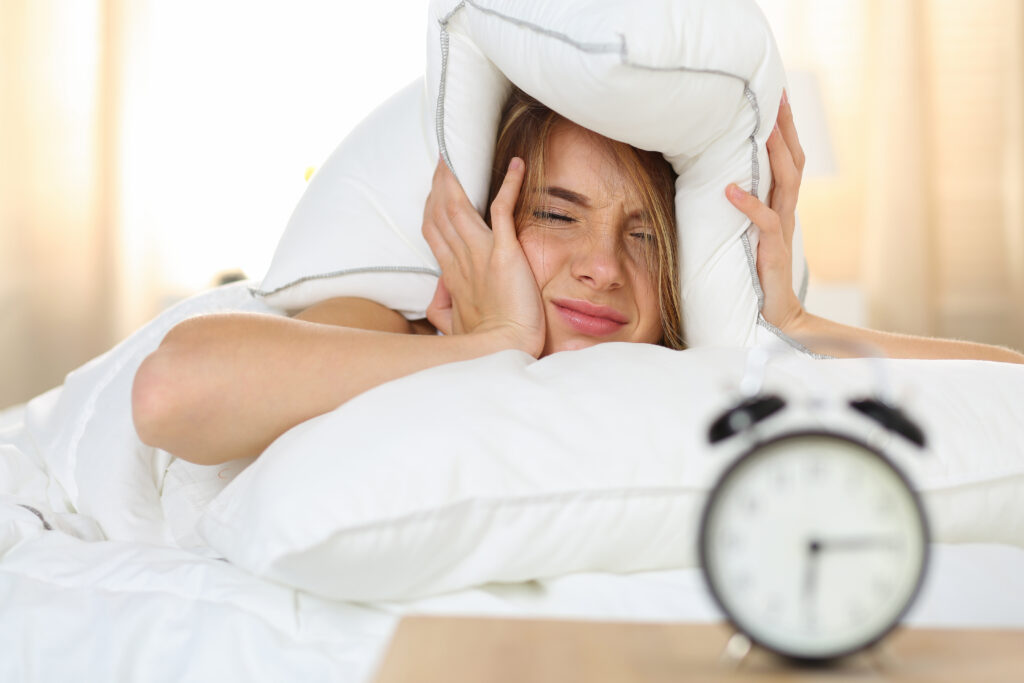 Young beautiful blonde woman lying in bed suffering from alarm clock sound covering head and ears with pillow making unpleasant face. Early wake up, not getting enough sleep, 