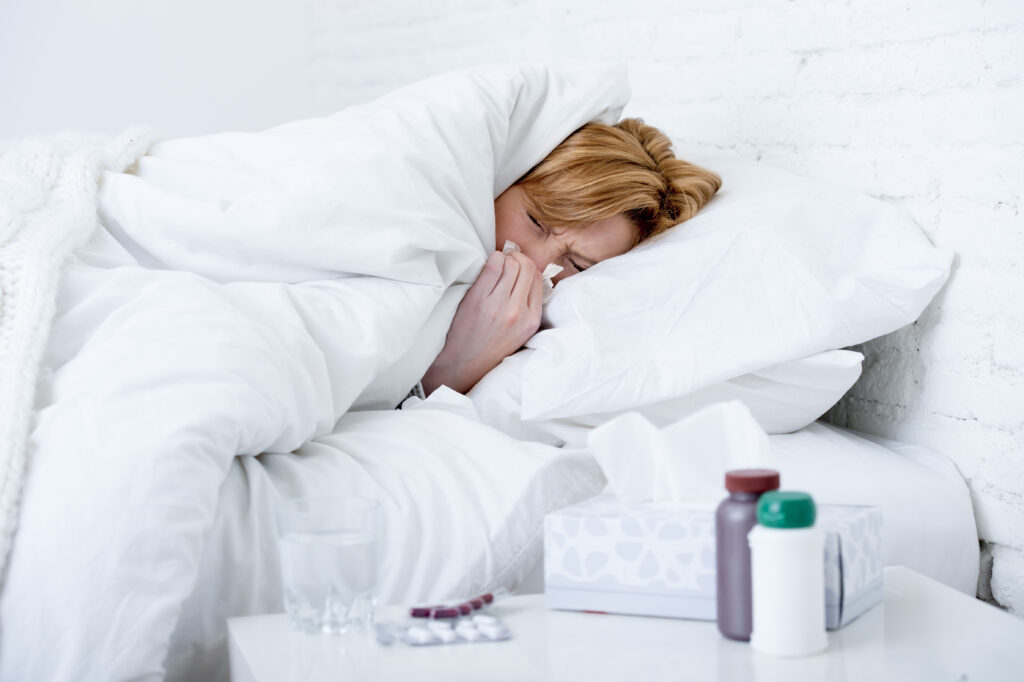 young sick woman with sneezing nose blowing in tissue lying on bed suffering winter cold and flu virus symptoms having medicines tablets and pills in illness and domestic health care concept; 