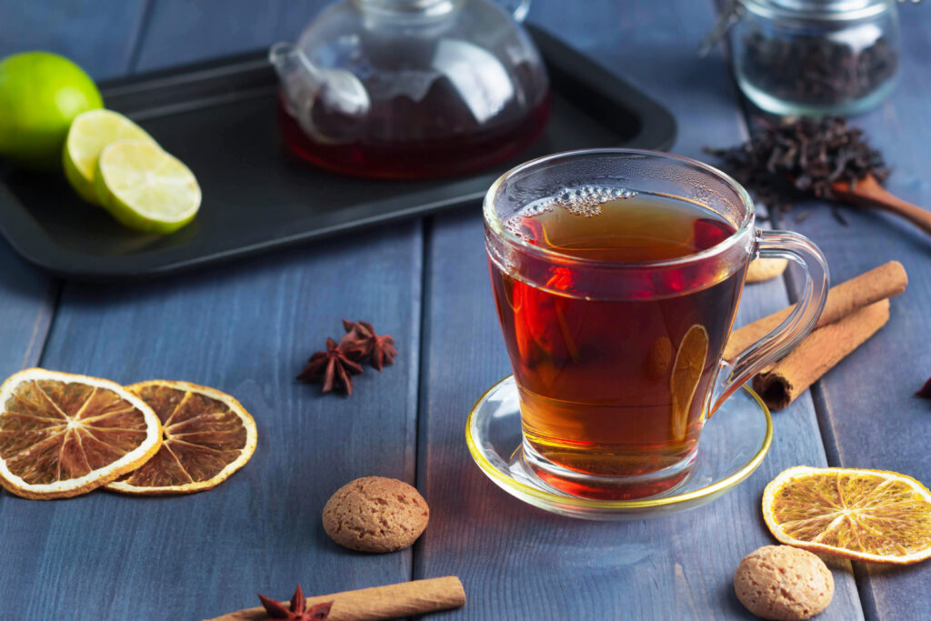 black tea in a transparent glass on a wooden table, next to a teapot, orange slices, cinnamon and cookies; 