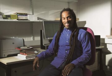 Benjamin Zephaniah, wearing a blue shirt, sits at a desk laden with books.