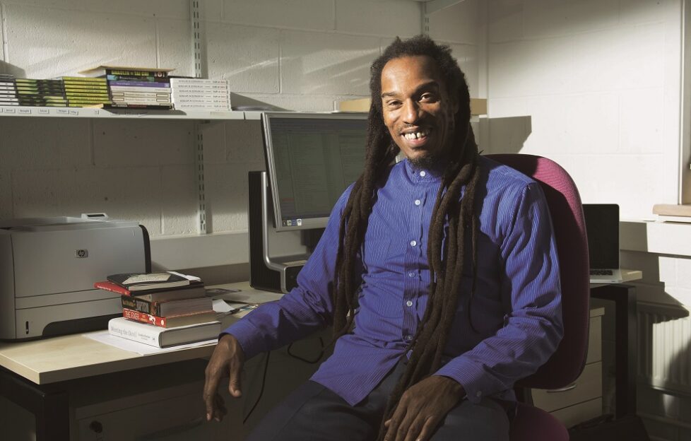 Benjamin Zephaniah, wearing a blue shirt, sits at a desk laden with books.