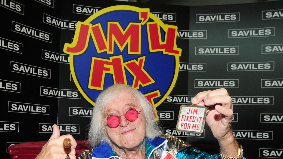 Jimmy Savile holds up Jim'll Fix It medal in front of TV show logo.