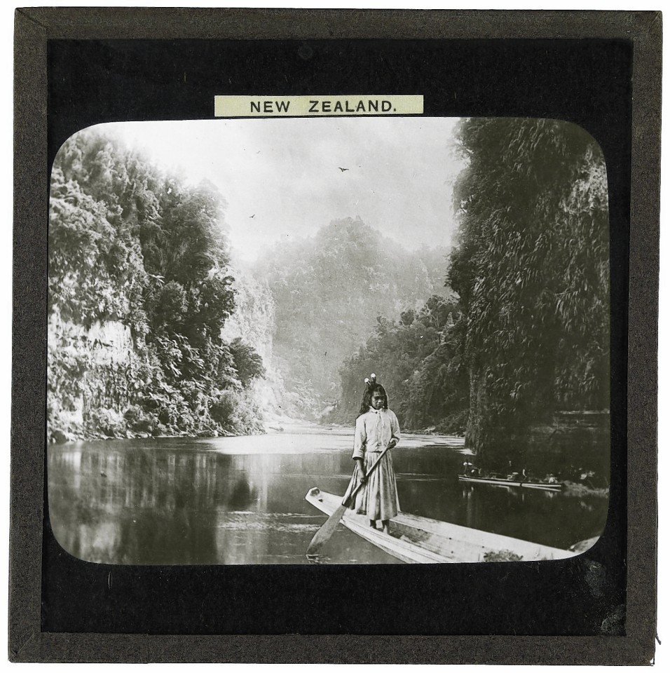 Photograph of a Maori girl standing on a canoe at the drop scene two miles up the Wanganui River in New Zealand in the late 19th or early 20th century. In the background there is another canoe, jungle and mountains.