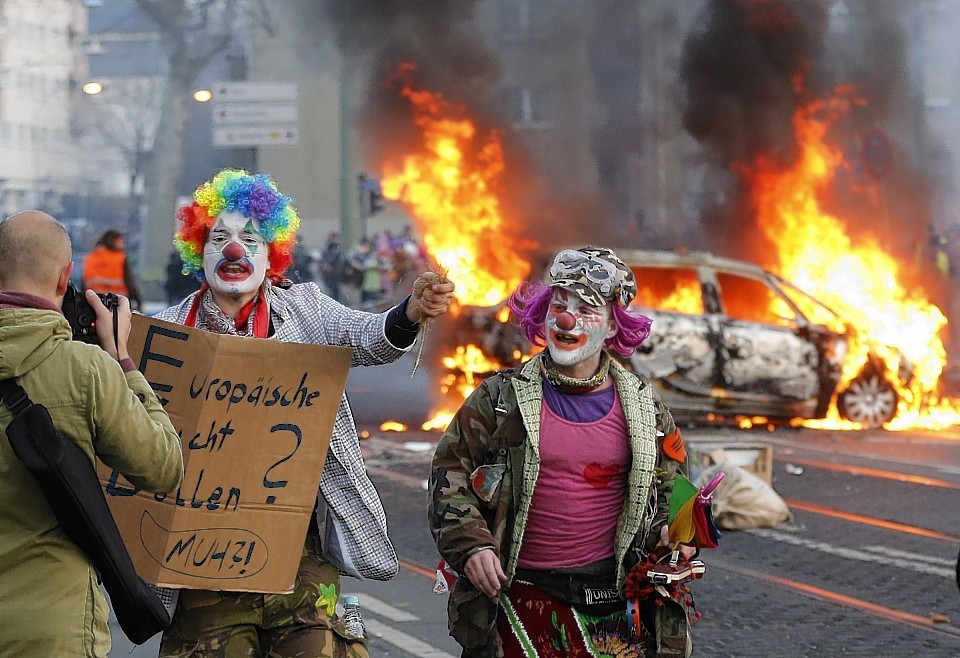 Demonstrators dressed as clowns pass by a burning police car