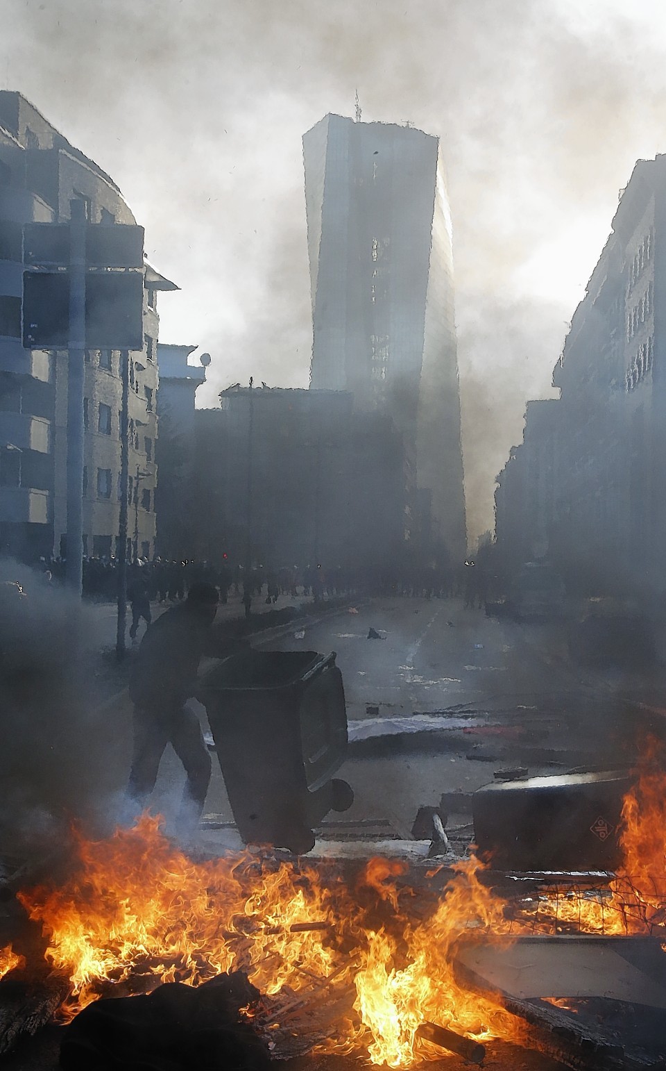 A protestor empties a trash bin as smoke billows over burning barricades in front of the new headquarters 