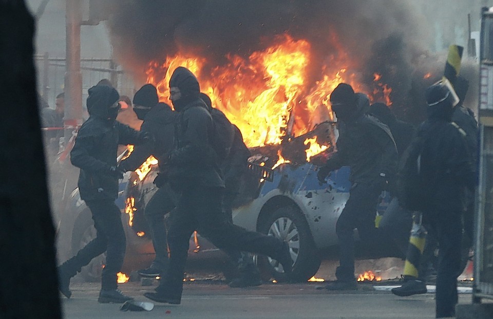 Masked demonstrators run past a burning police car 