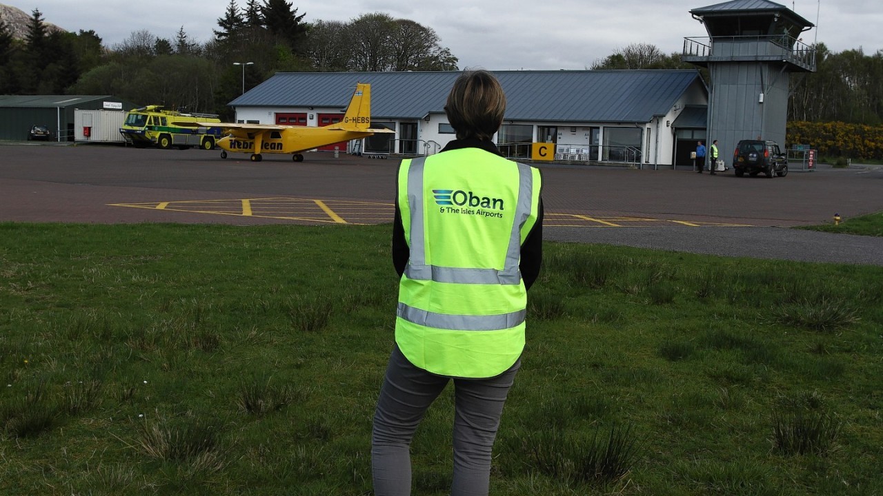 An Oban Airport worker stands in front of Oban Airport's building in a high viz vest. She is facing the Oban Airport building with a yellow aircraft on the runway. 