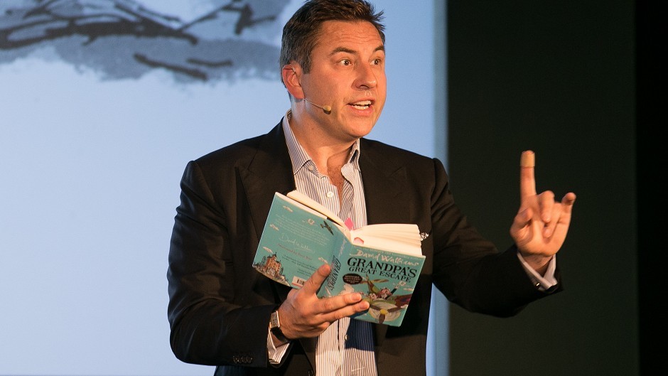 David Walliams standing on stage holding a book.