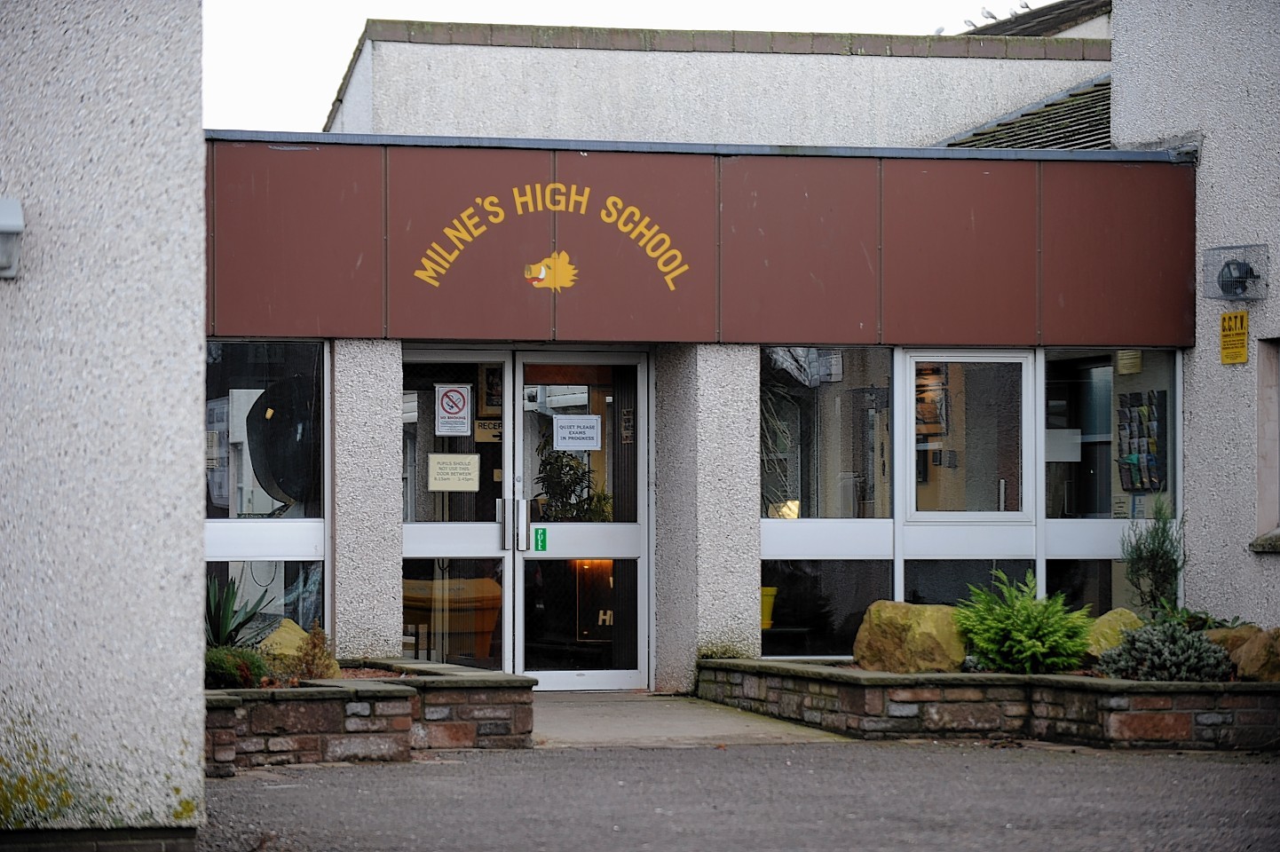 The exterior of Milne High School in Fochabers. Bedding plants are on either side of the door, with a bornw hand-painted sign above the door reading Milne High School. The building is a typical 1970/80s build, that looks a little run down.