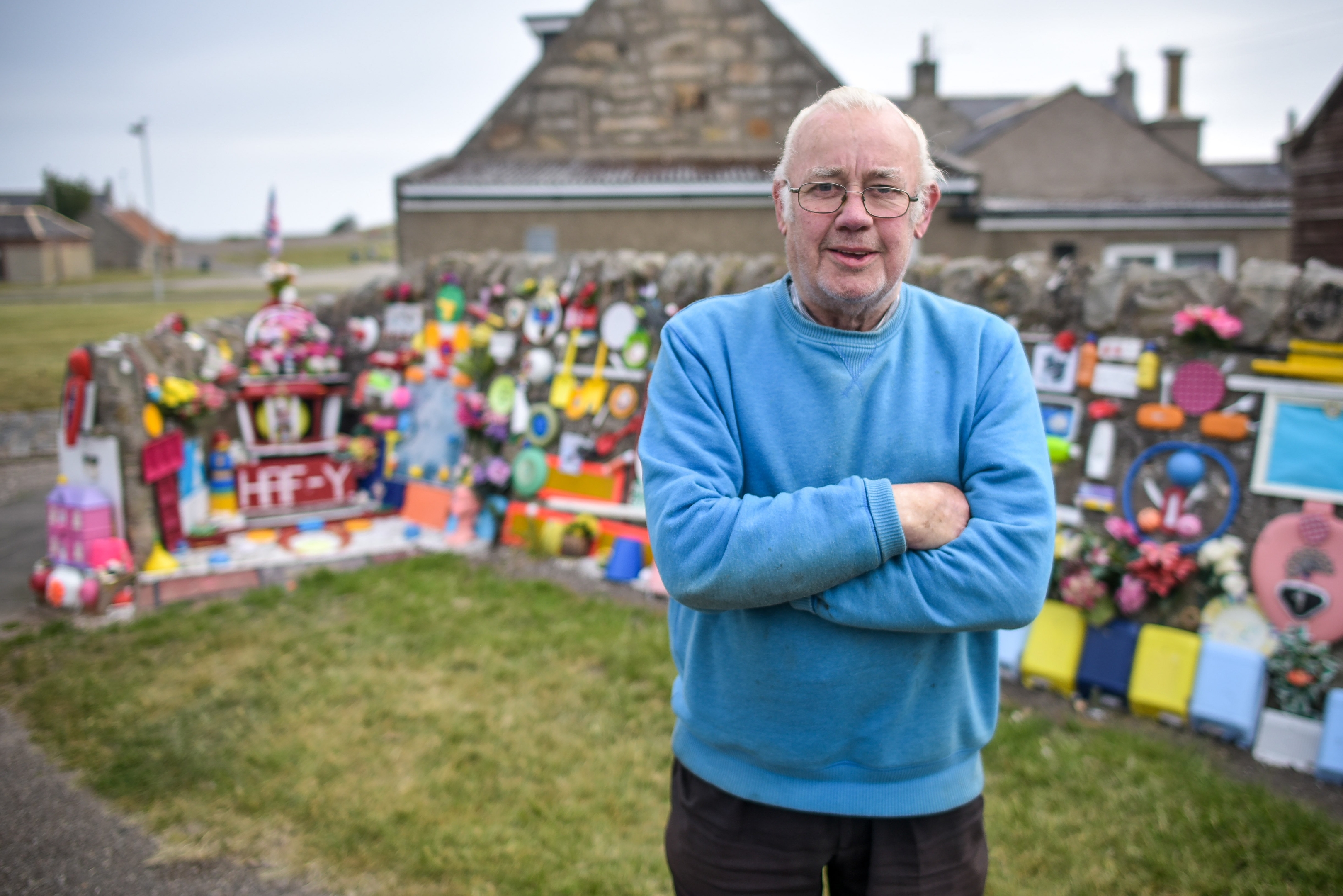 Charley Whyte, 76, with his mural on a wall in Lossiemouth, Moray on June 09 2016. Mr Whyte started the mural with just a chair in the corner as somewhere to sit during his evening walk. Since then he's added numerous other trinkets to the wall.  See Centre Press story CPLOO; A defiant retired plumber says he will not remove a bizarre mural he made from loo seats and toilet brushes. Charley Whyte, 76, started making the public artwork last August so that he could “brighten the place up”. And despite complaints from local residents, Charley said he won’t take down the mural in Lossiemouth, Moray. The mural, covered in random objects such as toilet brushes, loo seats and even Frisbees, stands at the entrance to Charley’s street. It is one of the first things drivers will see as they make their way into the town.