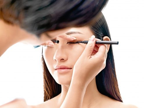 makeup artist working on eyeliner of a young asian model, isolated on white background.