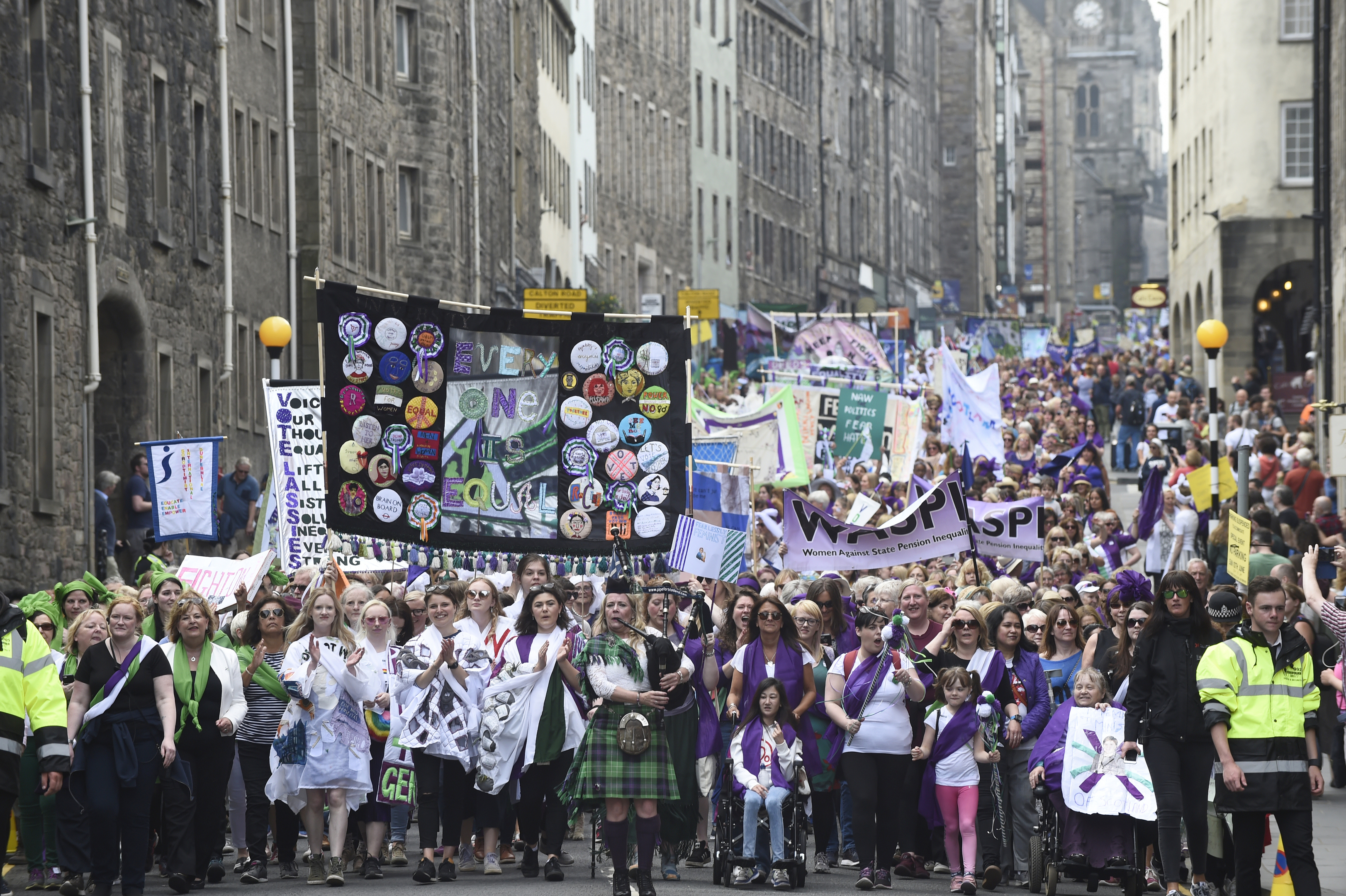 Women and girls walk together wearing scarves in either green, white or violet - the suffragette colours for Processions Edinburgh. The WASPI banner is clearly visible.
