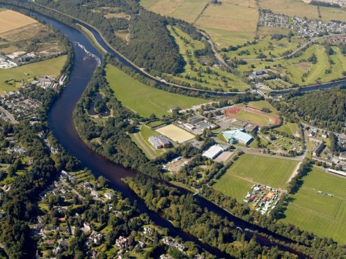 Bught Park, with its shinty pitches occupies the right foreground in this view, with the city's main leisure complex beyond.  This includes Queen's Park Sports Ground and Athletics Stadium; the ice rink, swimming pool and leisure centre, and the floral hall and botanic gardens. Torvean Golf Course lies beyond the Caledonian Canal in upper centre and right. A new road has proposed, which will link the Dores Road to the west (left) of the River Ness with the A82 Inverness to Fort William Road, on the western side of the River Ness and Caledonian Canal and this can be seen running though Torvean Golf Course in this view. The new road will cross the River Ness via a new bridge at the weir in the upper left of this view, and the Caledonian Canal via a swing bridge, which will open to permit traffic alond the busy canal.