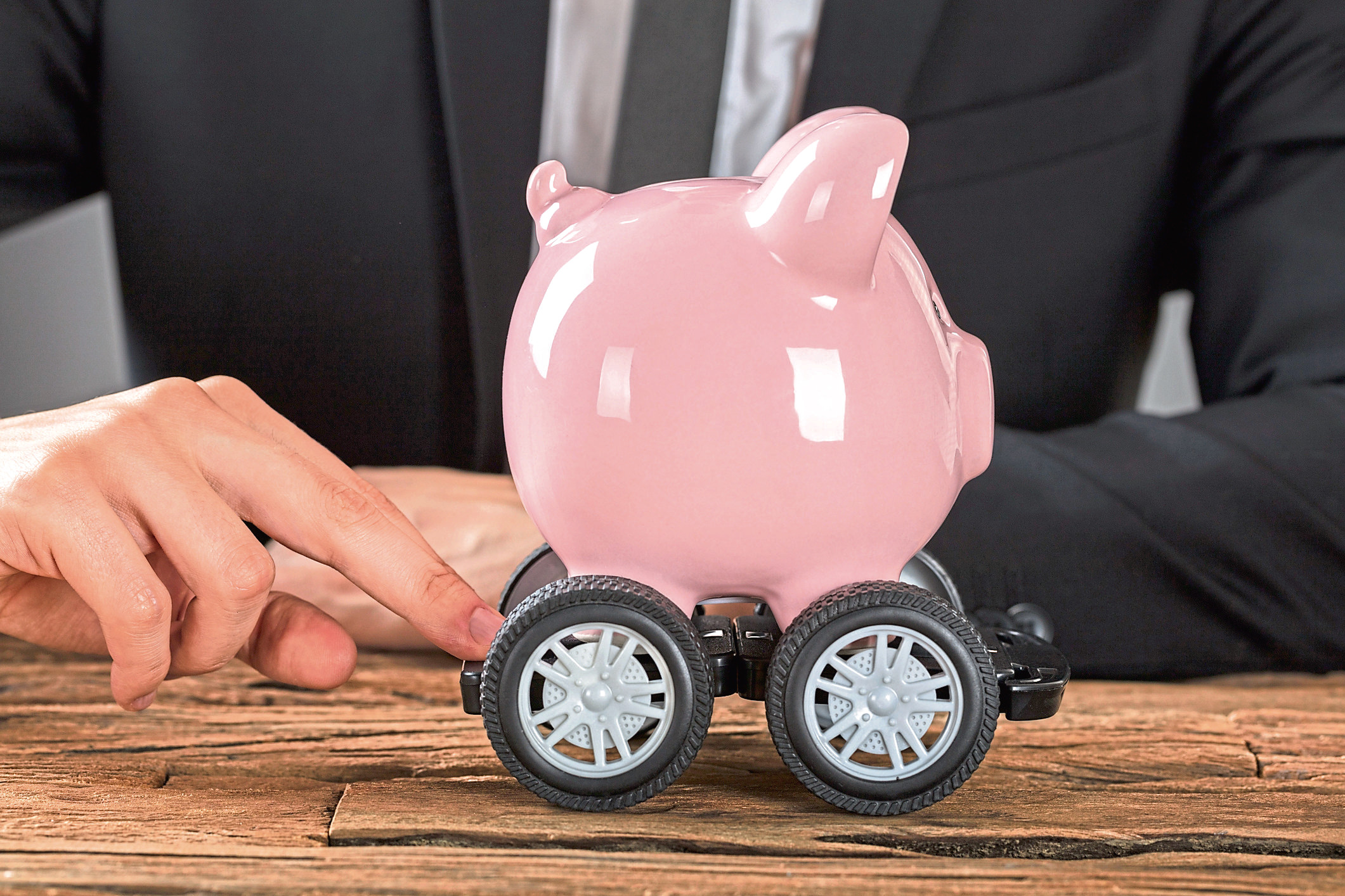 Close-up Of Businessperson Pushing Pink Piggy Bank On Wheels At Desk