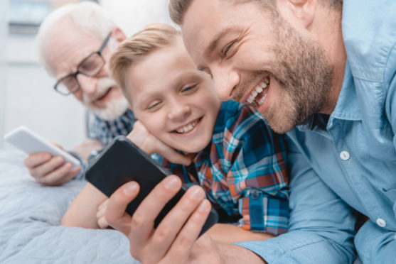 Little boy, his father and grandfather lying on bed with smartphones